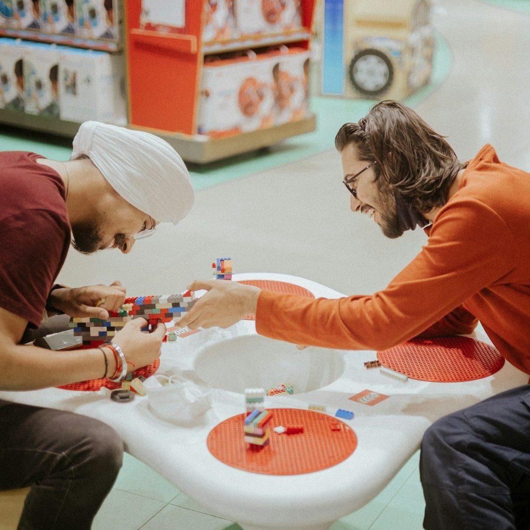 Deux personnes construisent ensemble avec des briques LEGO à une table de jeu blanche aux formes organiques, dotée de sections de plaques de base rouges intégrées. L'une porte un turban blanc et un t-shirt bordeaux, l'autre a les cheveux mi-longs, des lunettes et un pull orange — tous deux penchés en avant avec concentration et ce qui semble être un vrai plaisir. Le cadre ressemble à un magasin ou à un showroom LEGO, avec des présentoirs de produits visibles en arrière-plan.